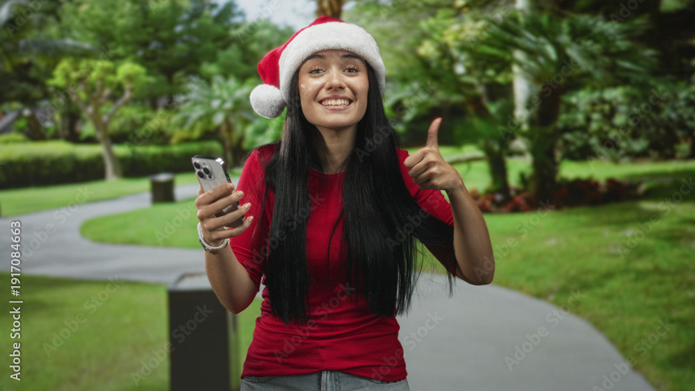Obraz premium Woman holding smartphone and waving hand while wearing a santa hat on a street pathway, smiling in surprise at her phone; holiday joy.