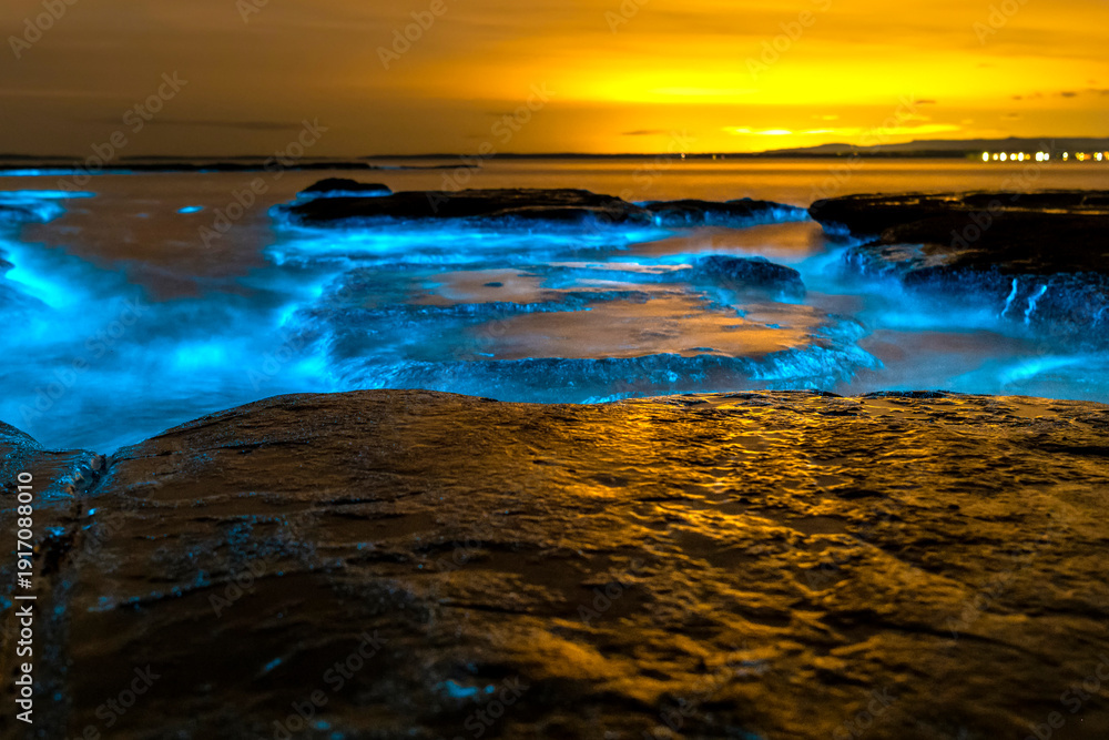 Naklejka premium Bioluminescent waves glowing blue around coastal rock platforms at night in Jervis Bay, New South Wales, Australia, with illuminated water and calm ocean conditions.