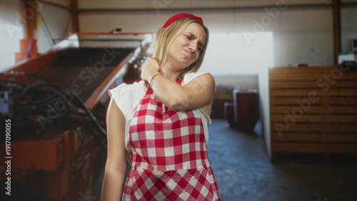 Woman cook in red check apron rubbing neck and grimacing inside a building with equipment and crates; discomfort.