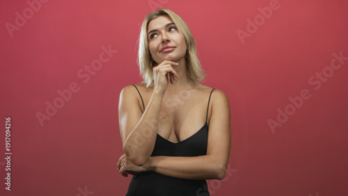 Young blonde woman touching chin with hand and crossed arms, wearing low-cut black tanktop against red studio backdrop; thoughtful contemplation.