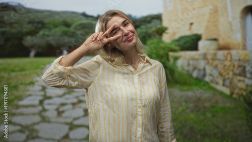 Young woman flashing a peace sign with her hand by her face in front of a stone building and a paved path; playful joy.