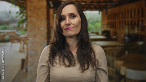 Wallpaper Mural Woman with long hair and beige sweater gestures questioningly against a rustic restaurant terrace backdrop with wooden furniture and soft natural lighting outdoors Torontodigital.ca