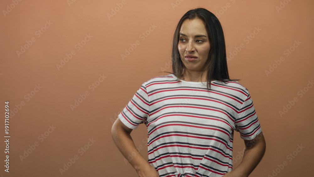 Obraz premium Woman grimacing in studio with peach background, wearing striped shirt and hands on hips showing a scrunched face; disgust discomfort.