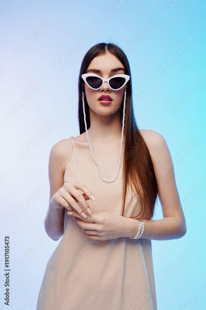 Fototapeta premium Young woman with long hair wearing white sunglasses and a beige dress poses confidently against a light blue background. Concept of beauty, fashion, and modern style.
