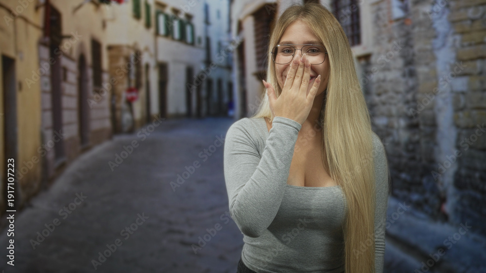 Fototapeta premium Young blonde woman with glasses covers mouth with hand on narrow cobblestone street; surprise reaction.