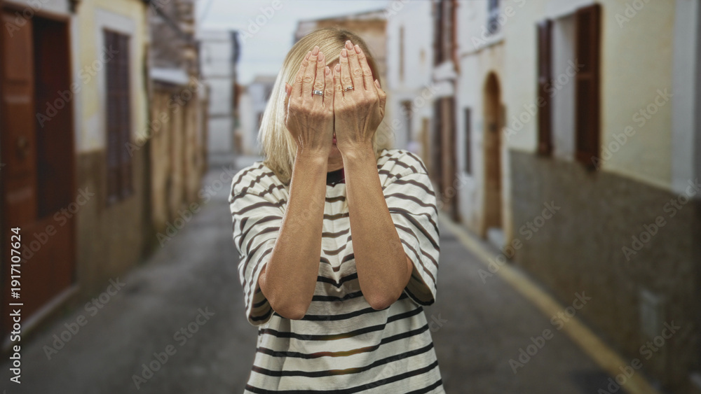 Fototapeta premium Woman covers face with hands and rings, wearing striped shirt, standing in a narrow street; playful curiosity.
