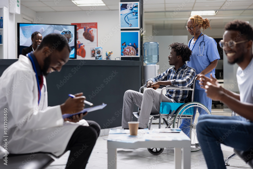 Obraz premium Nurse supports injured male patient seated in wheelchair while waiting for medical evaluation in hospital lobby. Doctor prepares documents and interacts with male visitor nearby in private clinic.