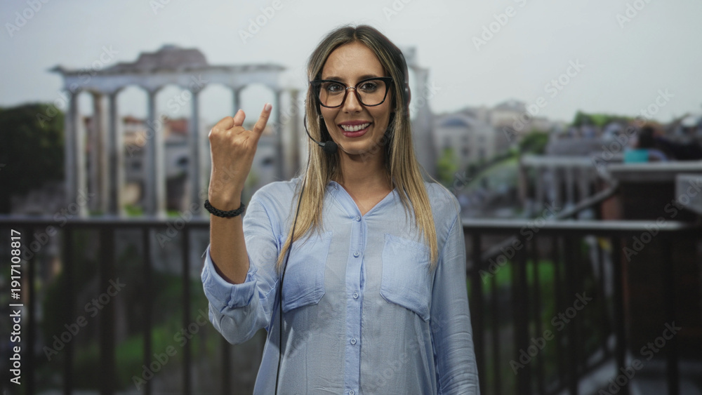 Fototapeta premium Woman wearing headset microphone with raised hand showing rock horns gesture at roman ruins building; customer service confidence.