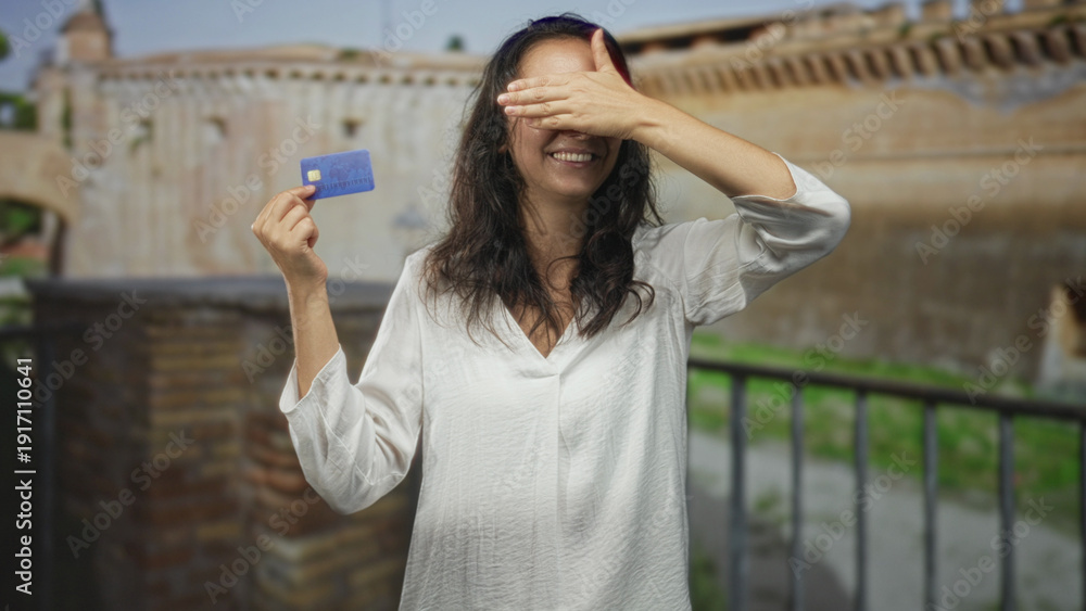 Fototapeta premium Woman holding blue creditcard between fingers, hand raised and smiling in front of a historic building; confidence ease leisure.