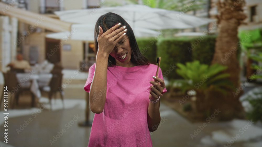 Fototapeta premium Young thai woman brunette holding a bamboo toothbrush and pressing hand to cheek for toothache in street cafe with umbrellas and tables; discomfort.