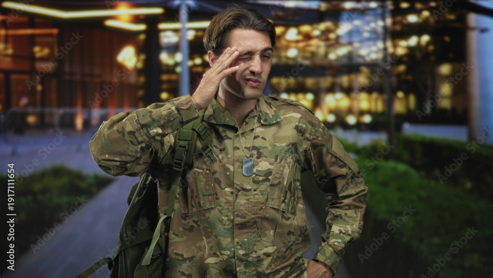 Obraz premium Man soldier in camouflage with backpack and dog tags, hand on forehead outside a lit building; duty fatigue.