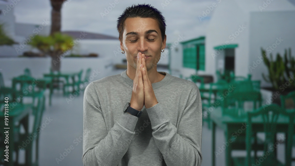 Fototapeta premium Young man in gray sweater stands smiling with hands together on a sunny outdoor terrace with green chairs and tables in the background of a bright restaurant setting