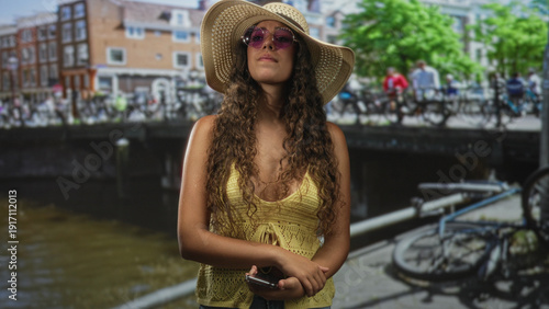 Hispanic teenage woman holding smartphone, wearing straw sunhat and pink sunglasses, looks up while standing on a street canal bridge; relaxed summer leisure.