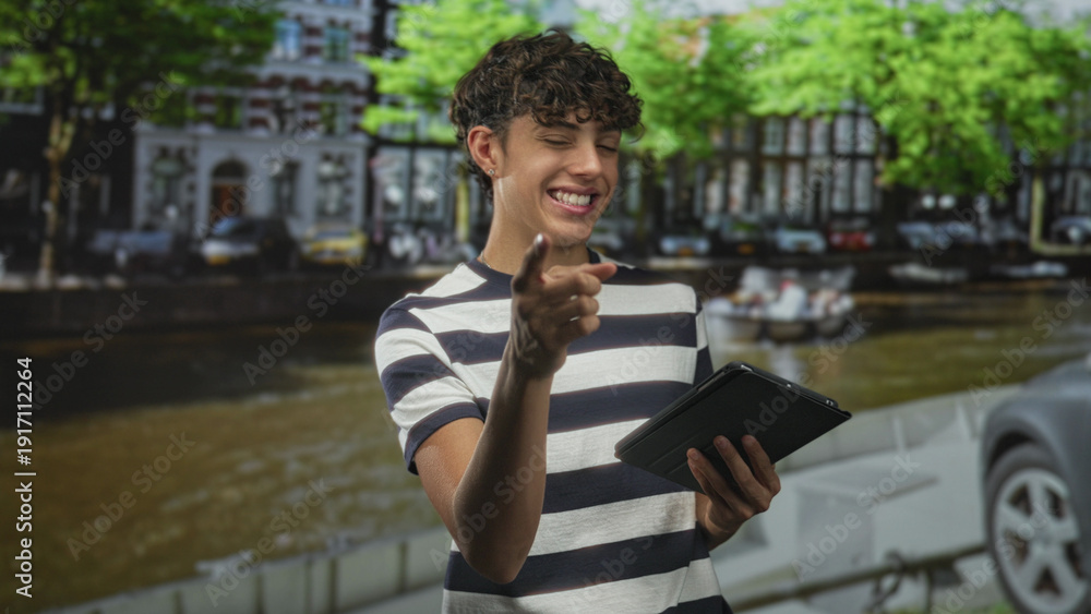 Fototapeta premium Boy in striped shirt points finger to tablet while holding the device on a street by a canal with parked cars and trees; playful curiosity.