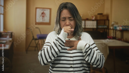 Schilderij op canvas Hispanic middleaged woman coughing into fist with hand on chest in clinic building during a medical exam; illness concern