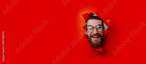 Fotografia Bearded Caucasian Man Bursting Through Red Paper With Joyful Grin, Round Glasses