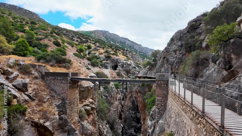 Handheld POV walking sideways right on the boardwalk of Caminito del Rey, revealing the deep gorge and a distant service bridge against the limestone cliffs.
