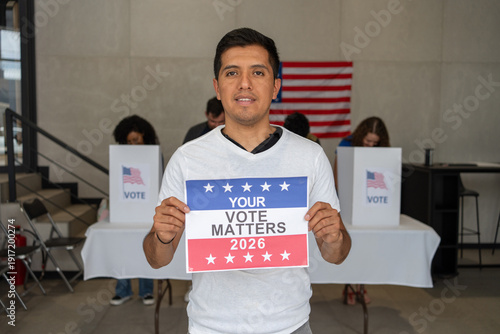 latino hispanic man holding a Your Vote Matters 2026 sign at a US election polling station. In the background, a diverse group of people votes in voting booths, democracy in America