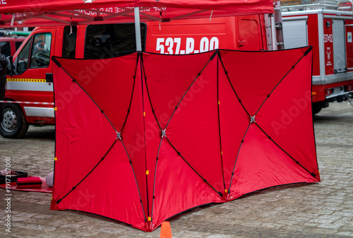 Red emergency incident screen shielding a medical rescue scene, mobile privacy barrier on cobblestones with volunteer fire department trucks and rescue equipment in the background.
