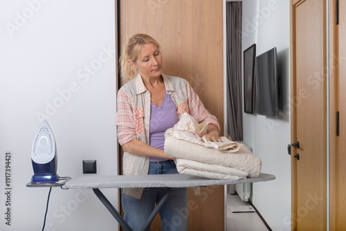 An adult woman is folding freshly ironed bed linen against a white wall with a wooden panel and a full-length mirror