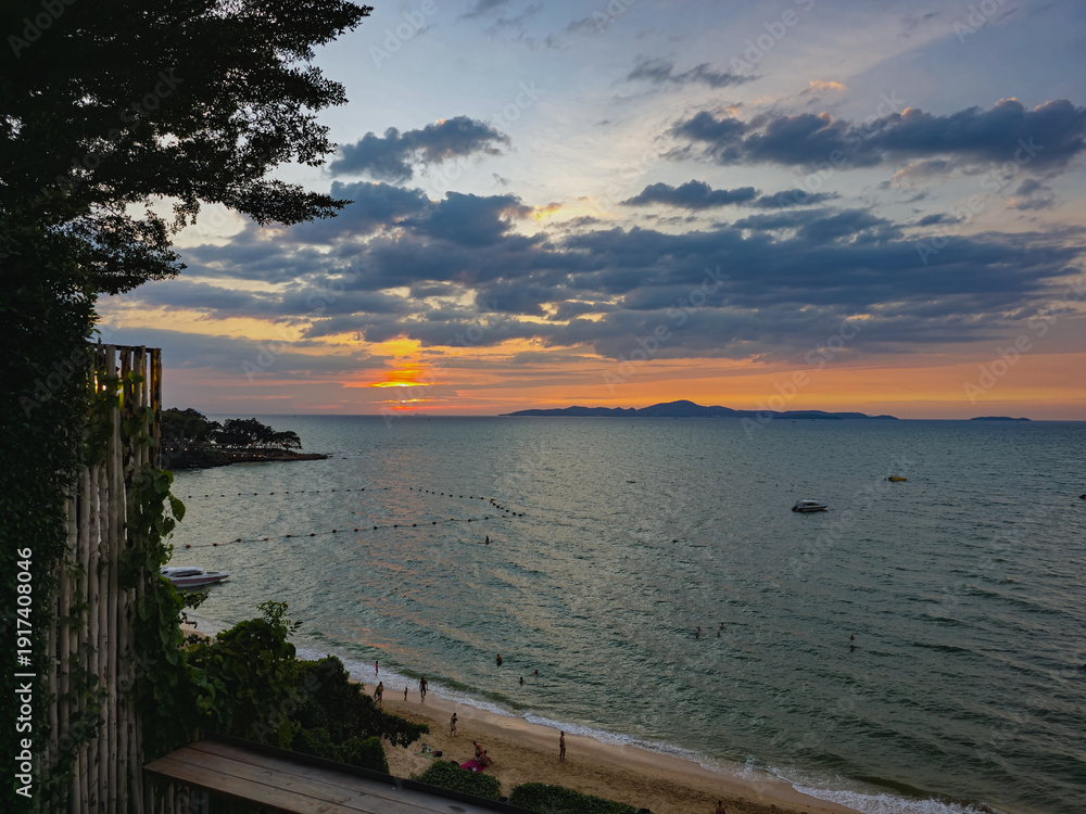Obraz premium A breathtaking sunset over a tropical bay with people relaxing on the beach and boats anchored in the calm sea under a dramatic cloudy sky