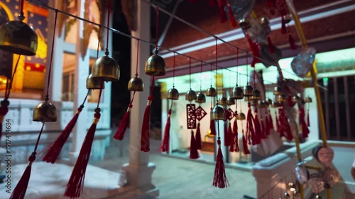 Row of bronze wind bells hanging in Thai Buddhist temple garden, Traditional sacred temple bells along railing with tropical foliage 4k