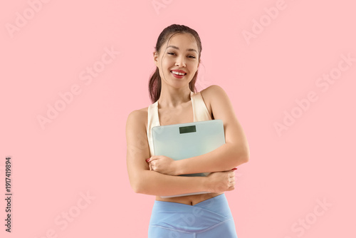 Sporty young Asian woman with weight scales on pink background. Diet concept