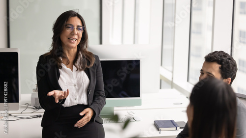 Mature 60s businesslady conducting informal meeting or briefing with team members, smiling, exchanging work-related ideas or thoughts, making decisions together, engaged in teamwork in coworking space