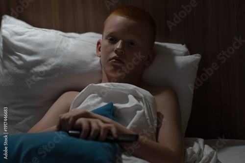 Obraz na plátně A red-haired boy lying in bed turns on his side, covers himself with a blanket and falls asleep in a hotel room after an interesting day