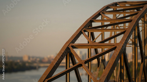 Rusty steel truss bridge arch detail at golden sunset with city skyline in background