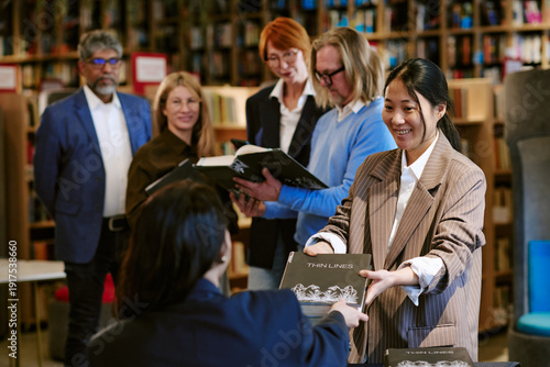 Fotografía Asian young adult woman handing book to seated female author while diverse group