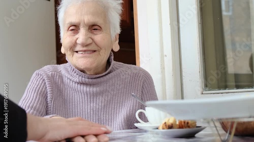 Female hand gently strokes hand of elderly woman sitting at table. Love and care for aging parents, guardianship of seniors and homeless elderly. Sweet gray haired grandmother with bright eyes.
