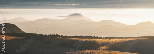 View of golden sunlight kissing the rolling hills and distant, mist-shrouded mountains crowned with a cloud-veiled peak, Mont-Dore, Auvergne-Rhone-Alpes, France.