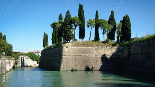 Alte Stadtmauern mit Zypressen bepflanzt und umgeben von einem Wasserkanal in der Nähe des Gardasees in Peschiera del Garda, Italien
