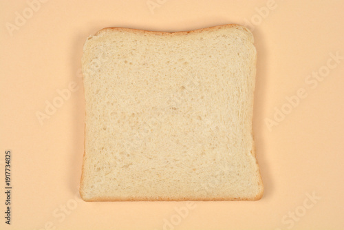 A single square slice of fresh white sandwich bread is centered on a plain light beige background shot from a top down perspective.