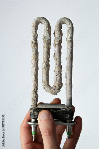 Hand holding used washing machine tubular electric heater covered in limescale.