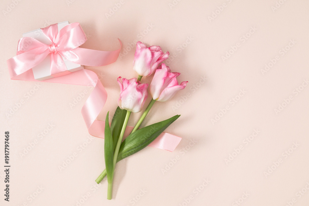 Obraz premium Pink tulip flowers and white gift box with silk ribbon on pastel beige background, top view flat lay with copy space. International Women's Day 8 March greeting card concept. Spring celebration.