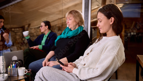 Young woman checking smartphone during informal office meeting. Concept of digital communication workflow, remote coordination, social media monitoring, modern hybrid workplace.