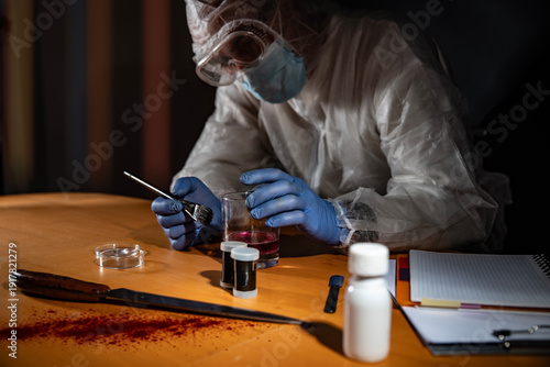Crime scene evidence processing on wooden desk: gloved hands use a brush to reveal latent prints on a drinking glass beside a knife with blood stains and sample vials.