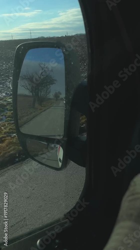 lonely tree reflected in side mirror, parked vehicle on shoulder, crisp light and faint frost on fields, quiet roadside atmosphere, soft glare across glass and contemplative pause