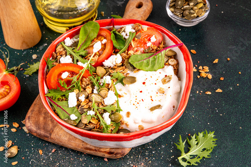 Savoury granola and yogurt breakfast bowl, balanced morning snack with greek yoghurt, tasty salted cheese granola, pumpkin seeds, nuts feta or tofu cheese, fresh vegetables and arugula