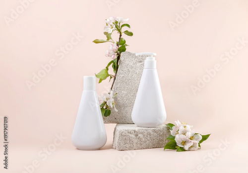 Two white cosmetic bottles on a stone podium surrounded by blooming apple twigs on a pastel background, natural skin care products.
