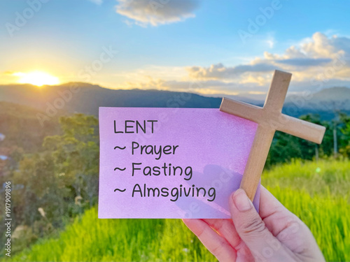 Billede på lærred Purple note with Lent, prayer, fasting, and almsgiving text held with wooden cross against sunset landscape, symbolizing the pillars of Lent season