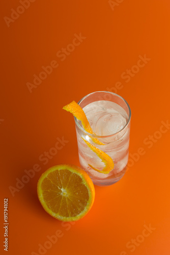 An overhead view of an iced coktail in a Hi Ball Galss with an orange twist and half an orange against an orange background