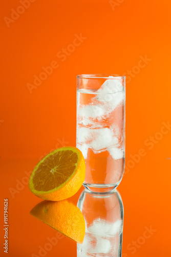 An iced cocktail in a HiBall glass with half an orange against an orange background with a relection of the glass and orange.
