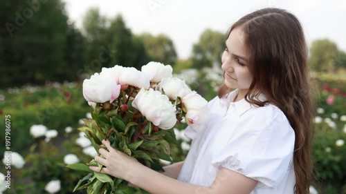 Young girl in white dress standing in peony field, holding pink flower bouquet. Smiling and smelling peonies. Summer nature background, romantic lifestyle concept, beauty and femininity. Copy space.