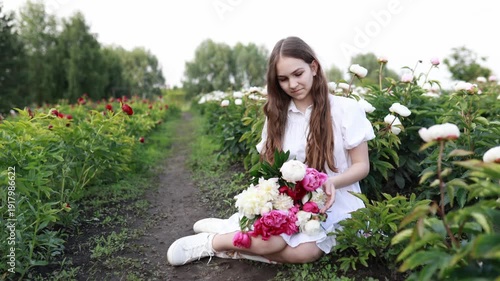 Young woman in white dress sitting in peony field, holding pink bouquet. Smiling, enjoying summer nature. Romantic lifestyle concept, beauty and femininity, soft sunlight. Copy space.