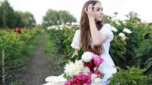 Young woman in white dress sitting in peony field, holding pink bouquet. Smiling, enjoying summer nature. Romantic lifestyle concept, beauty and femininity, soft sunlight. Copy space.