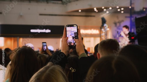 Young fans filming a concert on smartphones in a mall atrium. A huge crowd of teenagers gathered near the stage. Energetic audience enjoying a concert at a public event.