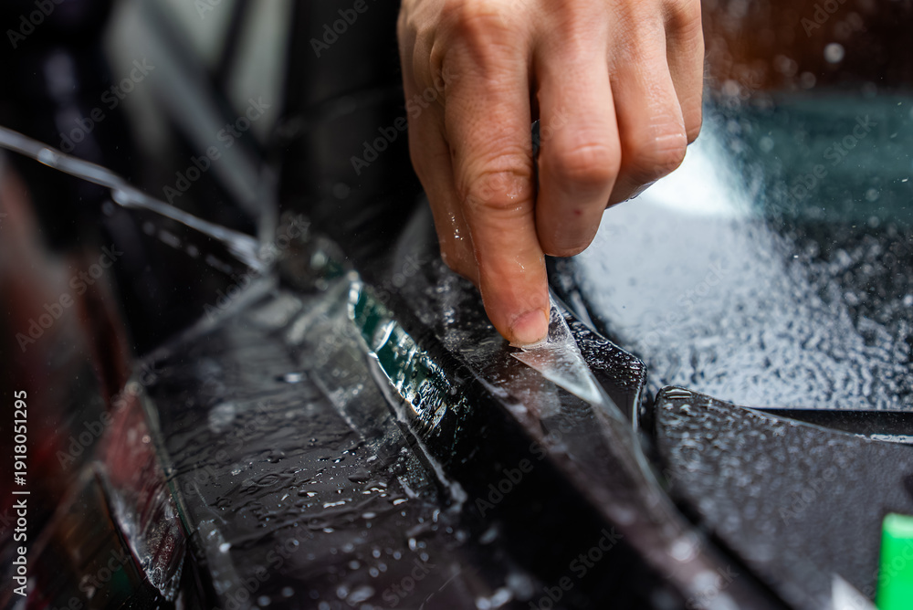 Fototapeta premium A technician peels clear protective film along a trim line on glossy black bodywork, with water beads and moody shallow depth lighting emphasizing precise craftsmanship.